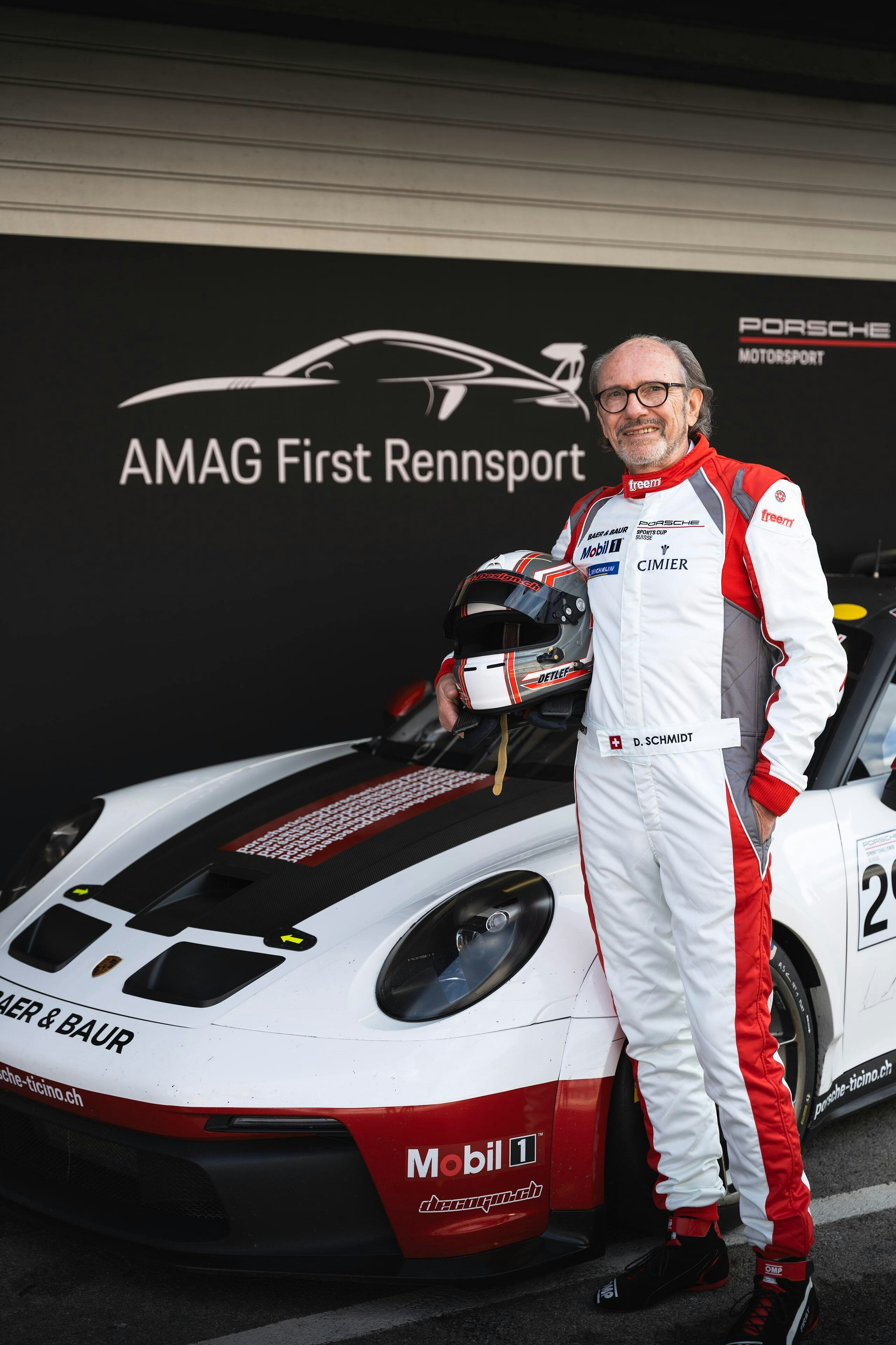 Detlef Schmidt im Rennazug mit Helm unter dem Arm neben einem Porsche-Rennwagen.